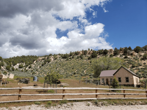 A rustic house and wooden fence in a grassy area, surrounded by hills and a partly cloudy sky.