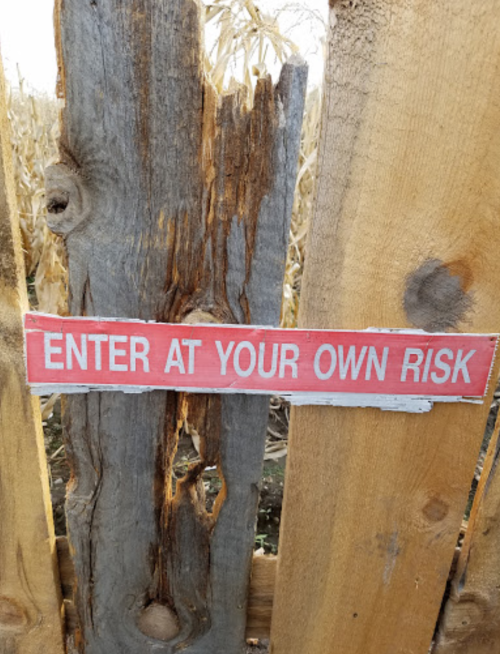 A weathered wooden sign reads "ENTER AT YOUR OWN RISK," attached to a rustic fence near a cornfield.