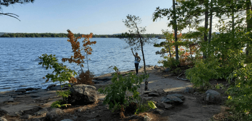 A person stands by a serene lake surrounded by trees and rocks, with colorful foliage in the background.