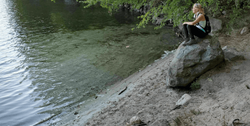 A person sits on a large rock by a calm lake, surrounded by greenery and sandy shore.