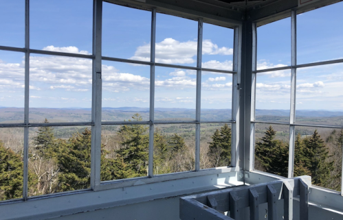 View from inside a lookout tower, featuring expansive mountains and a clear blue sky with scattered clouds.