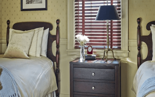 Cozy bedroom with two beds, a nightstand, a lamp, and a clock, featuring warm tones and natural light.
