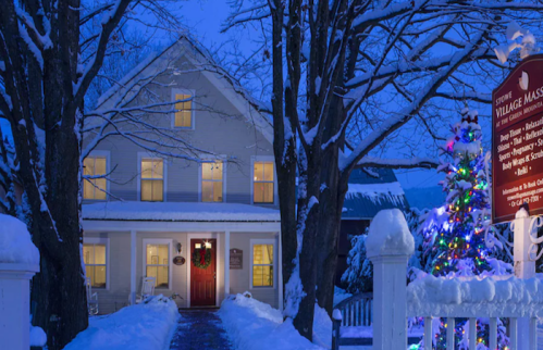 A snowy scene of a charming house at dusk, adorned with a festive wreath and a colorful Christmas tree nearby.