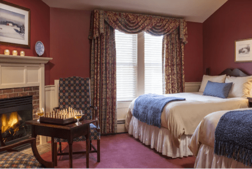 Cozy hotel room with two beds, a fireplace, and a chess set, featuring warm red walls and elegant curtains.