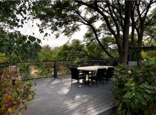 A wooden deck surrounded by trees, featuring a round table and black chairs, with greenery in the foreground.