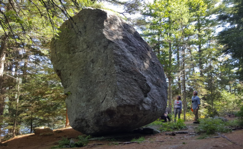 A large boulder stands in a forest, with two people nearby for scale among the trees.