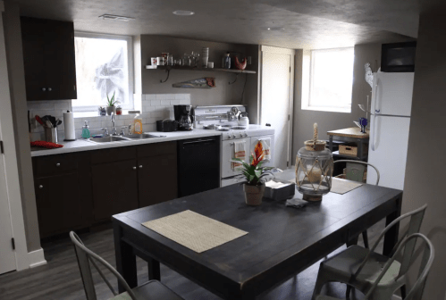 A modern kitchen with dark cabinets, a dining table, and large windows, featuring plants and kitchen appliances.