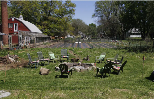 A circular seating area with green chairs surrounds a fire pit, set in a garden with rows of crops and a barn in the background.