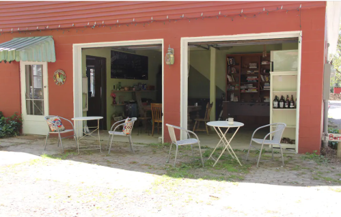 A cozy outdoor seating area with small tables and chairs in front of a red building with open doors revealing a warm interior.
