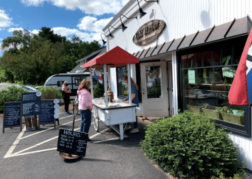 A quaint shop with a red awning, outdoor menu boards, and customers browsing on a sunny day.