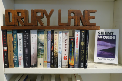A wooden sign reads "DRURY LANE" above a shelf filled with various books, including "SILENT WORDS" by Joan M. Drury.