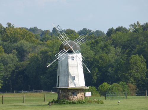 A white windmill with a stone base stands in a green field, surrounded by trees under a clear blue sky.