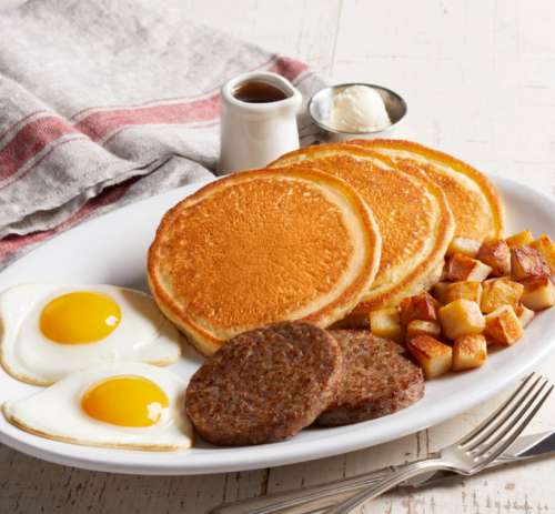 A plate of pancakes, fried eggs, sausage patties, and diced potatoes, served with syrup and butter on the side.