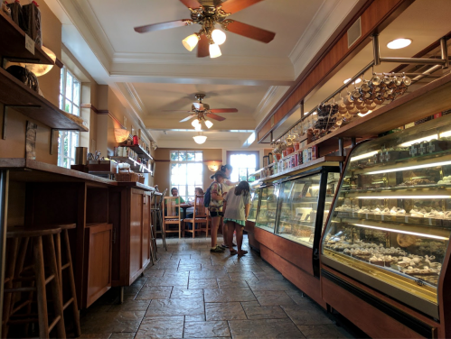 A cozy café interior with wooden shelves, a glass display case, and patrons seated at tables. Two people are chatting.