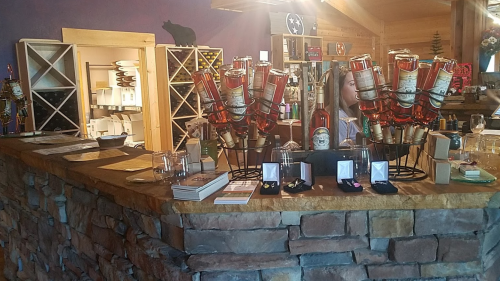 A rustic bar counter with bottles of liquor, glasses, and display items in a cozy, wooden shop setting.