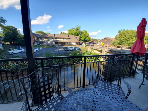 View from a patio with a black metal table and chairs, overlooking a creek and nearby buildings under a blue sky.