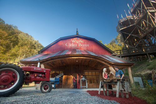A rustic cider barn with a red roof, a tractor in front, and people enjoying the outdoor space in a scenic setting.