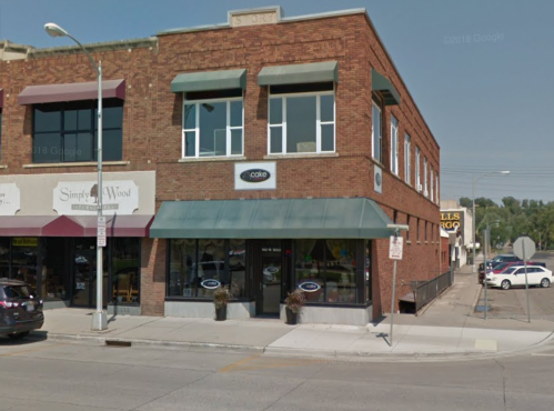 A brick building with large windows, featuring a green awning and storefronts on either side, located on a city street.