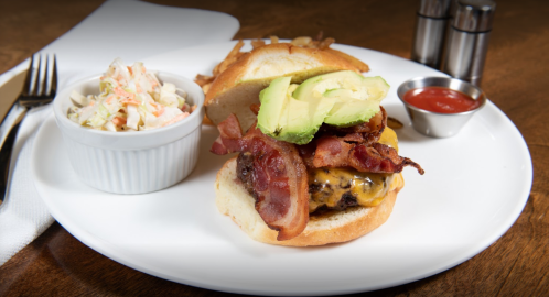 A juicy cheeseburger topped with bacon and avocado, served with coleslaw and a side of ketchup on a white plate.