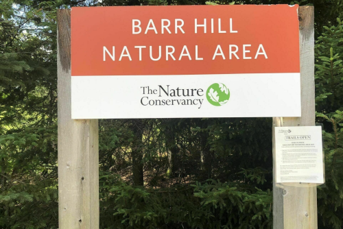 Sign for Barr Hill Natural Area by The Nature Conservancy, with information about open trails. Surrounded by greenery.
