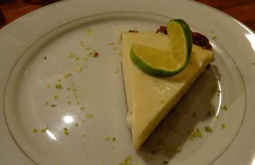 A slice of key lime pie on a white plate, garnished with a lime wedge and sprinkled with lime zest.
