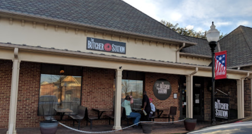 Exterior of The Butcher Station restaurant, featuring brick walls, outdoor seating, and an American flag banner.