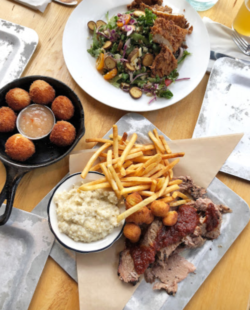 A wooden table with a plate of fries, brisket, hushpuppies, and grits, alongside a salad and a bowl of fried balls.