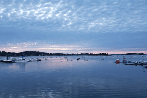 A serene harbor at dusk, with boats anchored in calm waters under a cloudy sky and soft pink horizon.