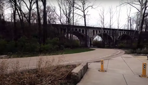 A winding path leads to a stone arch bridge surrounded by bare trees and greenery on a cloudy day.