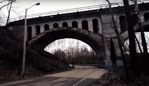 A large, arched stone bridge with multiple arches, surrounded by bare trees and a winding road beneath it.
