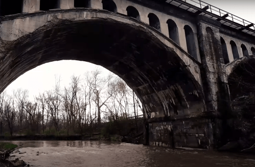 An old stone bridge arches over a river, surrounded by bare trees and a cloudy sky.