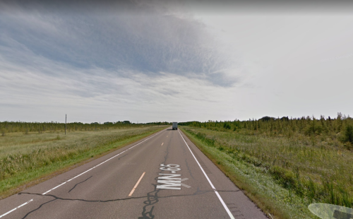 A long, empty road stretches through grassy fields under a cloudy sky, with a truck in the distance.
