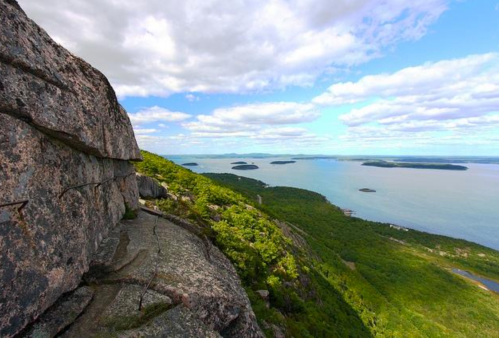 A scenic view from a rocky mountain overlooking a lush green landscape and a calm blue sea with islands in the distance.