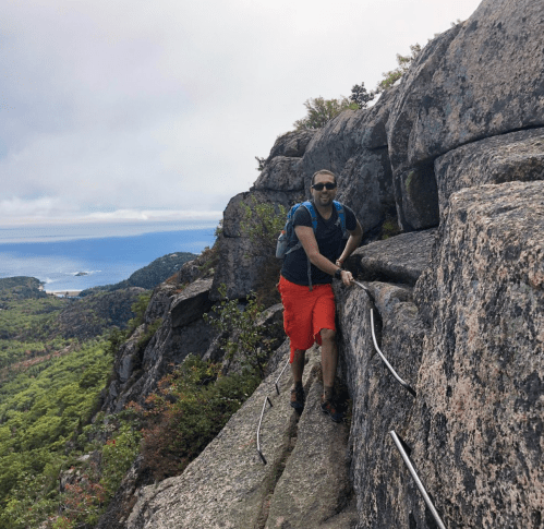 A hiker in orange shorts climbs rocky terrain with a scenic view of the ocean and greenery in the background.