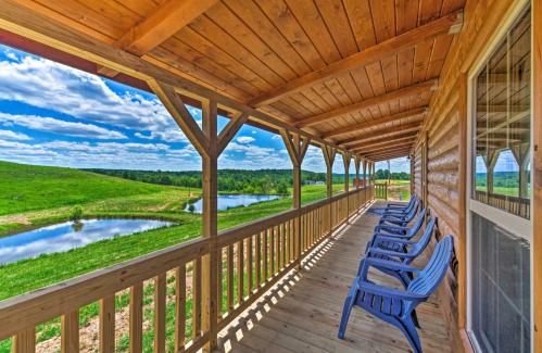 A wooden porch with blue chairs overlooks a lush green landscape and a serene pond under a bright, cloudy sky.