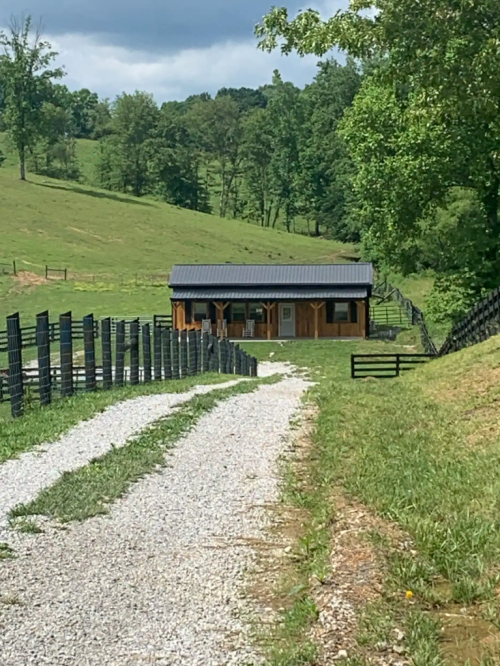 A gravel path leads to a wooden cabin surrounded by green hills and a black fence under a blue sky.