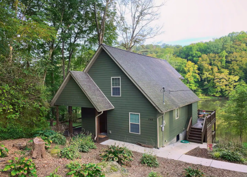 A green house surrounded by trees, with a deck overlooking a calm body of water and lush greenery in the background.