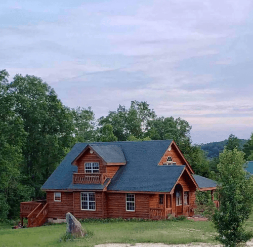 A charming wooden cabin surrounded by trees, featuring a sloped roof and a small balcony under a cloudy sky.