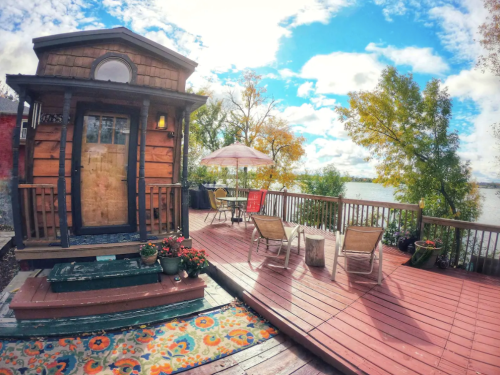 Cozy wooden cabin on a deck by the water, surrounded by trees and colorful autumn foliage under a bright sky.