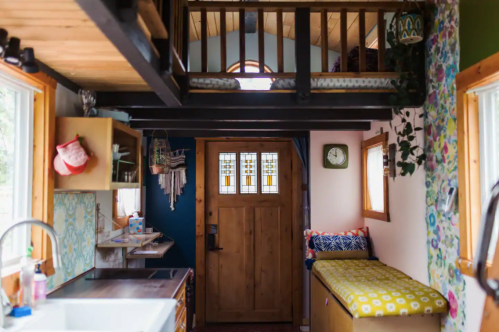 Cozy tiny home interior with a loft, wooden door, kitchen area, and colorful decor. Natural light fills the space.