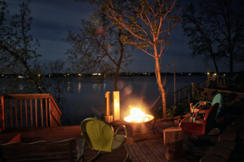 A cozy lakeside scene at night with a fire pit, colorful chairs, and trees reflecting on the water.