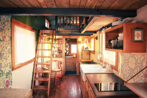 Cozy tiny house interior with a wooden staircase, kitchen, and lofted sleeping area, illuminated by warm sunlight.