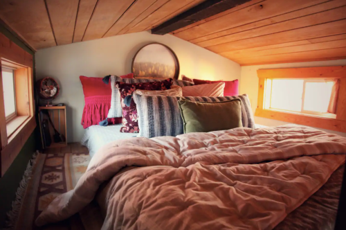 Cozy loft bedroom with a bed adorned in colorful pillows and blankets, featuring wooden beams and warm lighting.