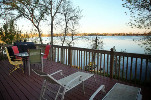 A serene lakeside view from a wooden deck with chairs and a table, surrounded by trees and calm water.