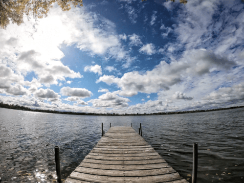 A wooden dock extends into a calm lake under a bright blue sky filled with fluffy clouds.