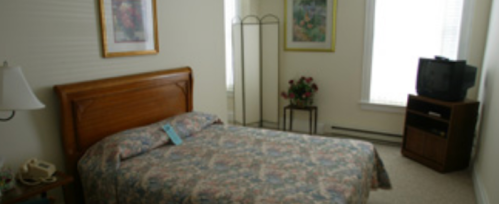 A simple bedroom with a floral bedspread, a wooden bed frame, a TV, and a small table with flowers.