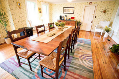 A cozy dining room with a wooden table, eight chairs, a colorful rug, and floral wallpaper.