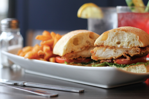 A close-up of a chicken sandwich with lettuce and tomato, served with curly fries and a drink in the background.