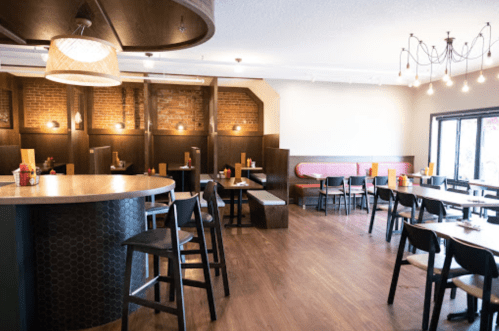 A modern restaurant interior featuring wooden floors, brick walls, and a mix of seating options at tables and a bar.