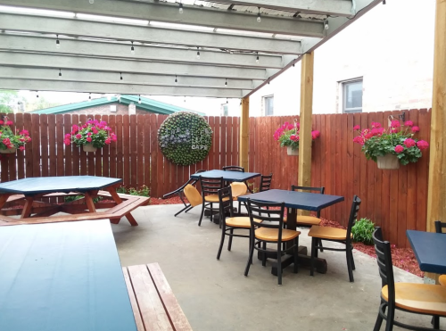 Outdoor café seating area with wooden tables, chairs, and vibrant flower planters against a wooden fence.